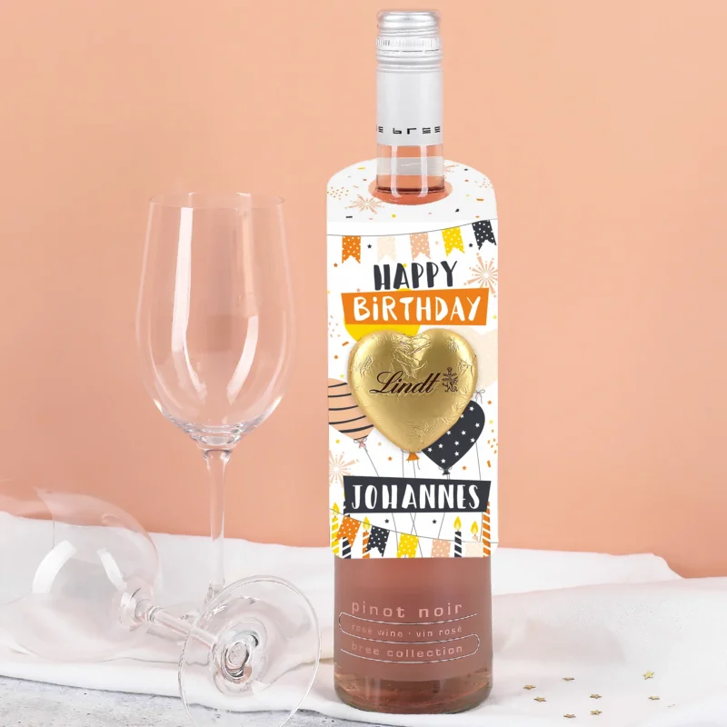 Flaschenanhänger mit „Happy Birthday“-Design in Weiß, Orange und Schwarz, goldfarbenes Lindt Schokoladenherz und personalisierter Name – an Flasche befestigt.